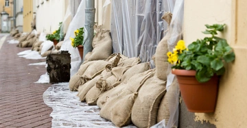 Aufgestapelte Sandsäcke vor dem Garagentor eines städtischen Reihenhauses. Unter und über den Säcken ragt eine transparente Plastikfolie hervor, die das Garagentor abdeckt. Links und rechts davon jeweils ein Blumentopf mit gelben Primeln.