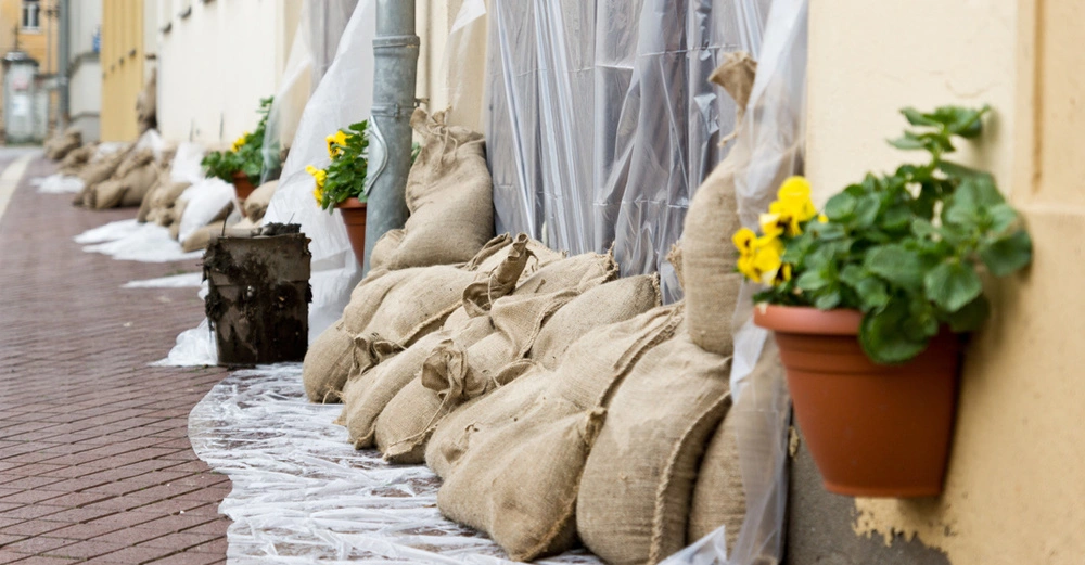 Aufgestapelte Sandsäcke vor dem Garagentor eines städtischen Reihenhauses. Unter und über den Säcken ragt eine transparente Plastikfolie hervor, die das Garagentor abdeckt. Links und rechts davon jeweils ein Blumentopf mit gelben Primeln.