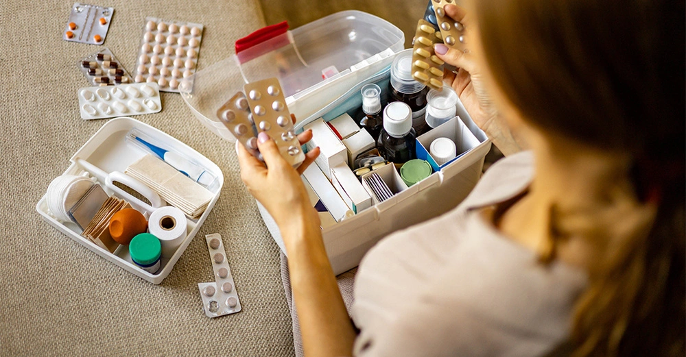 Person sortiert verschiedene Medikamente in Pillenpackungen und Fläschchen in einer großen weißen Box auf einem braunen Stoff. Daneben befindet sich eine kleinere Box mit Verbandsmaterial.