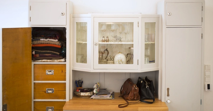 Galerie-Bild 4: Ein weißer Schrank enthält Kleidung Gläser und Dekorationsgegenstände auf einem Holzregal in einem hellen Zimmer mit beige Wänden Ein schwarzer Hut liegt oben auf dem Schrank