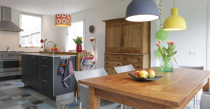 Galerie-Bild 4: Holztisch steht in einer modernen Küche bunte Kronleuchter hängen darüber Schüsseln mit Obst Vase mit Tulpen auf dem Tisch schwarze Arbeitsplatte bunte Fliesen Holzkasten Fenster und Stühle