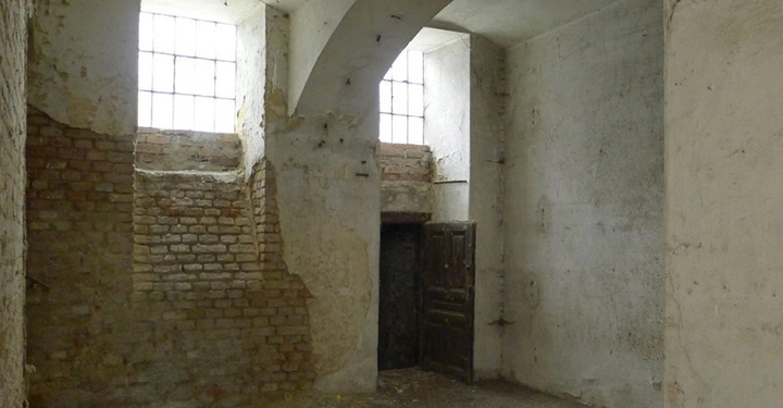 Galerie-Bild 8: Verlassener Raum mit bröckelnden Ziegelwänden und schmutzigen Böden große weiße Gewölbedecke zwei vergitterte Fenster und eine braune Holztür links und hinten unverputzte Wände allgemein verfallen