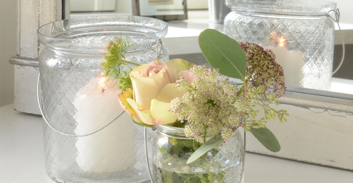 Galerie-Bild 9: Ein Glasbehälter enthält eine brennende weiße Kerze. Ein anderer Glasbehälter daneben hält Blumen. Im Hintergrund sind Spiegel und Fenster zu sehen, die die Szenerie reflektieren.