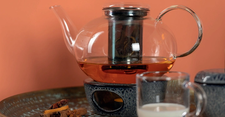 Glasteekanne mit Tee zieht auf einem schwarzen Untersetzer mit einem roten Hintergrund Ein Glas Milch und Gewürze wie Zimt und Sternanis sind auf einem silbernen Tablett arrangiert