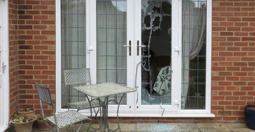 Ein zerbrochenes Fenster in einer weißen Terrassentür zeigt eine beschädigte Glasscheibe und zerbrochenes Glas auf dem Boden davor Ein Metalltisch mit zwei Stühlen steht auf einem Steinboden daneben