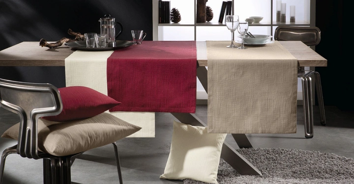 Ein Tisch mit einer roten Tischdecke und Geschirr steht in einem modern eingerichteten Raum. Dahinter befinden sich ein Regal mit Dekorationen und eine Metallschale. Kissen liegen auf Stühlen und dem Boden.