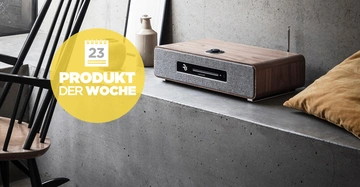 Holzlautsprecher spielt Musik auf einer Betonplatte neben einer braunen Vase und zwei gerahmten Bildern Text bei gelbem Kreis Kalender-Symbol 23 PRODUKT DER WOCHE dunkler Stuhl und Kissen in der Nähe