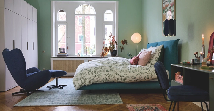 Ein Kingsize-Bett mit gemusterter Bettwäsche steht in einem hellen Schlafzimmer mit grün gestrichenen Wänden neben einer Lampe und Pflanzen vor einem großen Fenster mit Blick auf Bäume und Gebäude. Ein blauer Sessel und ein Schreibtisch stehen im Raum. Text im Bild: Flower Market Bologna.