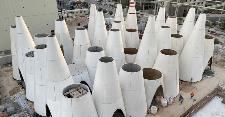 Galerie-Bild 7: Mehrere große, weiße, kegelförmige Strukturen stehen auf einer Baustelle. Arbeiter in orangefarbener Sicherheitskleidung gehen zwischen den Kegeln umher, umgeben von Gebäuden und Baugeräten.