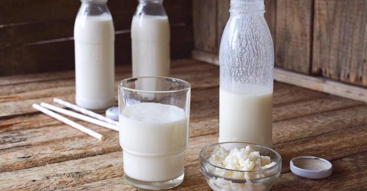 Galerie-Bild 4: Ein Glas Kefir steht auf einem Holztisch neben einem Glas mit Kefirkörnern und drei Flaschen Kefir im Hintergrund in einem rustikalen Raum.