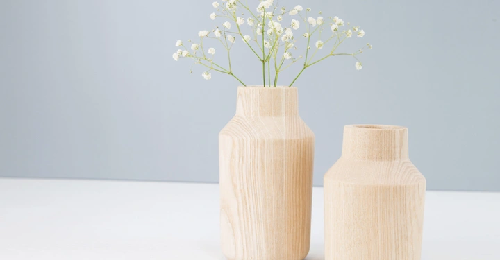 Galerie-Bild 9: Zwei Holzkannen stehen auf einer weißen Oberfläche in einem minimalistischen Raum. Eine Kanne enthält kleine weiße Blumen. Die Umgebung ist schlicht und hell.