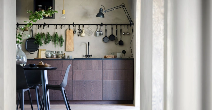 Dunkler Holztisch mit dunkelgrauem Stuhl steht in einer minimalistischen Küche mit hölzernen Unterschränken. Küchengeräte und Kräuter hängen an einer Wandstange, beleuchtet von einer Wandleuchte.