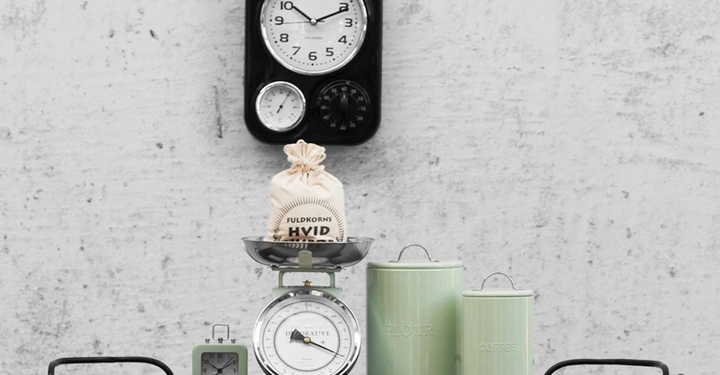 Galerie-Bild 12: Eine klassische Uhr zeigt die Zeit an einer Wand an. Darunter stehen eine Waage mit einem Mehlsack sowie zwei Vorratsdosen und eine kleine Uhr auf einem Regal. Text: FULDKORNS HVID FLOUR COFFEE DECORATIVE