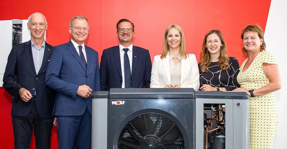 3 Männer in Anzug und 3 Frauen in Businesskleidung stehen lächelnd nebeneinander auf einer knallroten Bühne mit rotem Hintergrund. Vor ihnen eine Wärmepumpe von WOLF Österreich. 