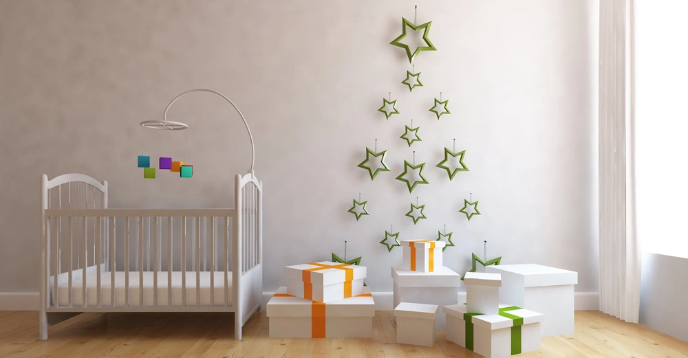 Ein weißes Babybett steht in einem hellen Raum mit Holzboden, an der Wand sind grüne Sterne als Baumform aufgehängt, daneben mehrere Geschenke mit orangefarbenen und grünen Schleifen.