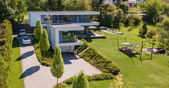 Ein modernes zweistöckiges Haus steht auf einem großen grünen Rasen mit einem Pool im Hintergrund. Zwei Autos parken auf der Auffahrt neben dem Haus.