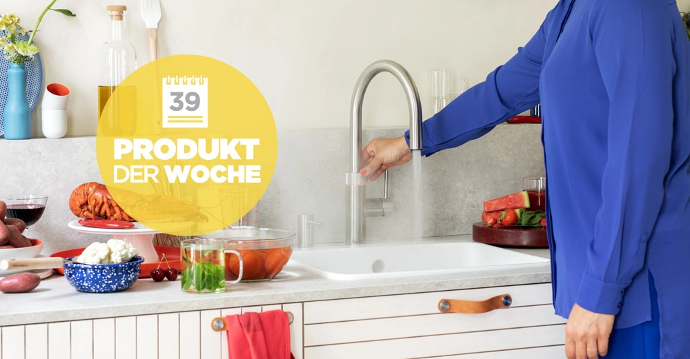 Person aktiviert Wasserhahn in einer Küche mit Arbeitsplatte voller frischer Zutaten wie Gemüse und Obst. Ein gelber Kreis mit der Aufschrift 39 PRODUKT DER WOCHE ist im Vordergrund.