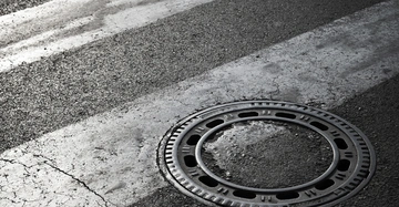 Gullydeckel liegt auf einer Straße mit weißen Zebrastreifen. Die Straße zeigt Abnutzungsspuren und Risse im Asphalt.