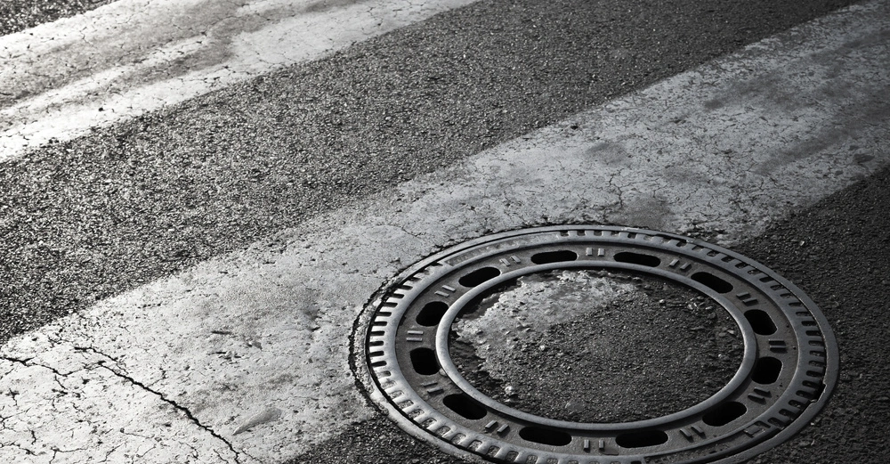 Gullydeckel liegt auf einer Straße mit weißen Zebrastreifen. Die Straße zeigt Abnutzungsspuren und Risse im Asphalt.