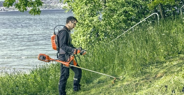 Eine Person trimmt das Gras mit einer Motorsense am steilen Ufer eines Sees, umgeben von dichtem grünem Laub.