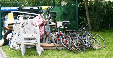 Plastikstühle Fernsehsessel Fahrräder und andere Haushaltsgegenstände liegen stapelweise auf einer Wiese in einem umzäunten Bereich in der Nähe von Mülltonnen und Bäumen