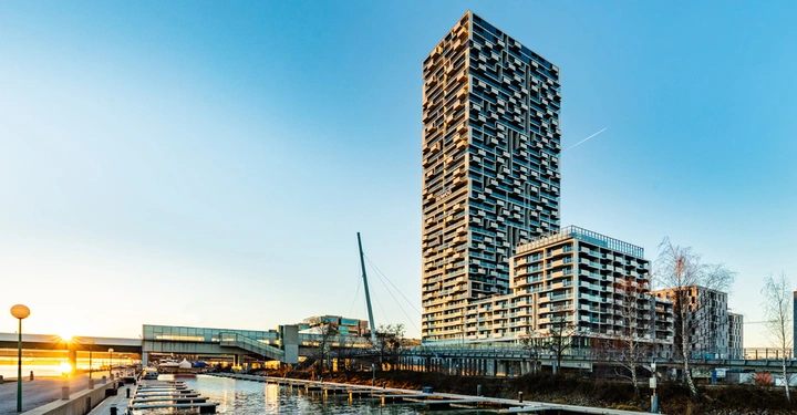 Ein hoher Wolkenkratzer mit geometrischem Design steht neben einem Wasserkanal bei Sonnenuntergang. In der Nähe befinden sich ein Steg mit kleinen Booten und moderne Gebäude. Text: BUWOG.