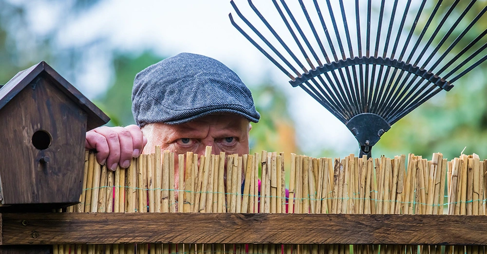 Ein Nachbar sieht böse über den Zaun an der Grundstücksgrenze mit einem Laubrechen in der Hand.