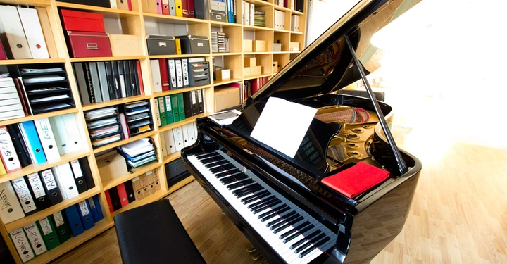 Galerie-Bild 1: Ein schwarzer Flügel mit geöffnetem Deckel steht spielbereit auf einem hellen Holzboden in einem Raum, der von deckenhohen Regalen mit bunten Ordnern umgeben ist.