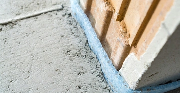 Nahaufnahme eines frischen Zementestrichs, der mit einem blauen Randdämmstreifen von der Ziegelwand getrennt ist