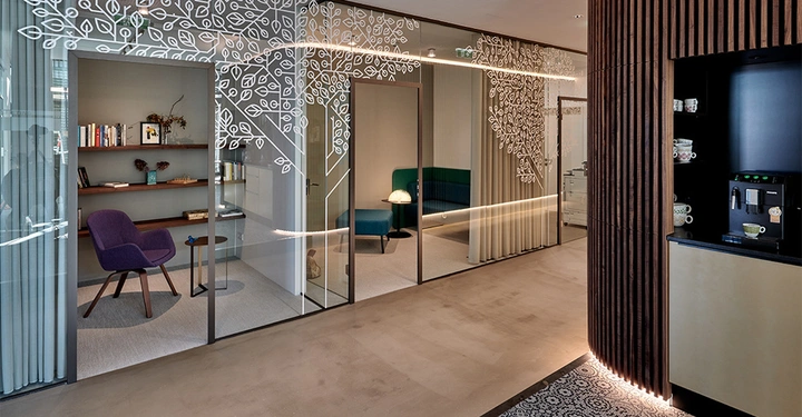 Lila Stuhl steht in einem gemütlichen Arbeitszimmer mit Regalen und Büchern an der Wand an einer verglasten Bürowand mit weißen Blattmustern entlang eines modernen Büroflurs mit Kaffeemaschine.