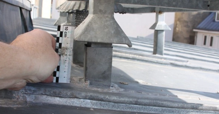 Galerie-Bild 19: Hand hält ein Lineal mit einer 10 cm-Skala nahe an einem Metallbauteil auf einem Dach in einer städtischen Umgebung. Das Dach ist metallisch und hat verschiedene Strukturelemente.