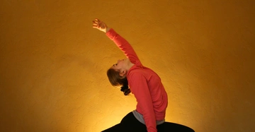 Eine Person macht eine Yoga-Pose in einem Raum mit gelber Wand. Sie trägt ein rosa Sweatshirt und schwarze Leggings während sie sich seitlich nach hinten streckt.