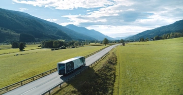 Ein Lastwagen fährt auf einer Landstraße durch eine grüne ländliche Umgebung mit Hügeln und Bergen im Hintergrund. Auf dem Lastwagen steht THE MAGIC OF INNOVATION.