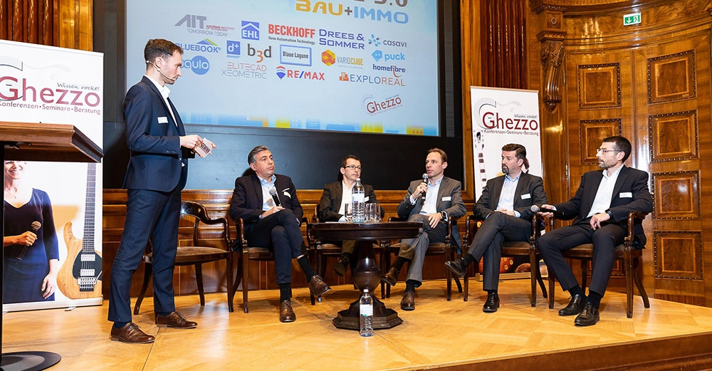 Fünf Männer sitzen auf Stühlen und unterhalten sich auf einer Konferenzbühne Ein Mann steht und hält ein Mikrofon Im Hintergrund ist eine Projektion mit verschiedenen Firmennamen zu sehen Texte: Ghezzo Wissen rockt Konferenzen Seminare Beratung