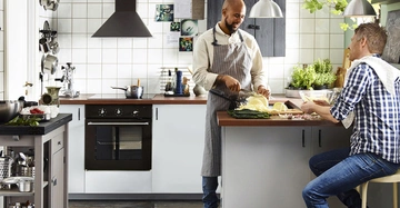 Zwei Männer unterhalten sich in einer modernen Küche während einer von ihnen Gemüse schneidet. Die Küche hat weiße Fliesenwände, eine zentrale Arbeitsplatte und verschiedene Küchenutensilien an der Wand.