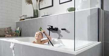 Kind badet in der Badewanne mit Schaum spielt mit Duschschlauch graue Fliesen weißer Rand Regale und Handtücher links Dekorationen oben gerahmte Bilder im Hintergrund.