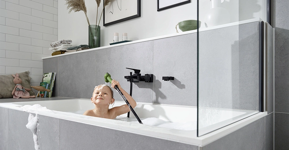 Kind badet in der Badewanne mit Schaum spielt mit Duschschlauch graue Fliesen weißer Rand Regale und Handtücher links Dekorationen oben gerahmte Bilder im Hintergrund.