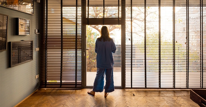 Frau steht mit Rücken zum Fotografen und blickt aus Ganzglasfensterfront mit Innenjalousien, angenehmes Licht im Raum.