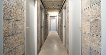 Keller eines Wohngebäudes, schmaler langer Flur mit jeweils 3 Kellertüren links und rechts und einer Tür an der Stirnseite. Wand, Boden und Türen in weiß, hellbraun und beige.