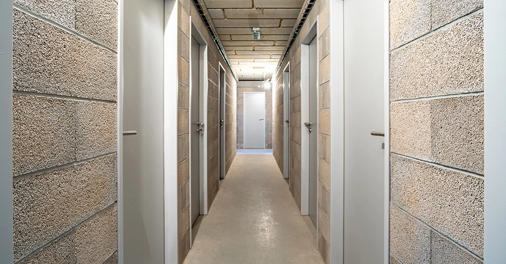 Keller eines Wohngebäudes, schmaler langer Flur mit jeweils 3 Kellertüren links und rechts und einer Tür an der Stirnseite. Wand, Boden und Türen in weiß, hellbraun und beige.