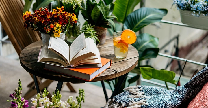 Auf einem runden Holztisch liegt ein geöffnetes Buch auf einem geschlossenen Buch Umliegende Gegenstände sind ein Glas Orangensaft bunte Blumen und grüne Pflanzen Der Kontext zeigt einen gemütlichen Balkon
