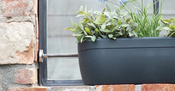 Grauer Blumenkasten mit grünen Pflanzen und blauen Blüten ist am Fenster montiert auf einer Ziegelmauer.