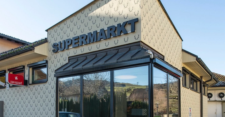 Galerie-Bild 2: Gebäude mit dem Schriftzug SUPERMARKT; links ein Schild ÖSTERREICHISCHE LOTTERIEN; Hintergrund ländliche Umgebung mit Hügeln und Himmel. Fassadenmuster bestehen aus kleinen schwarzen Hirschgeweihen.