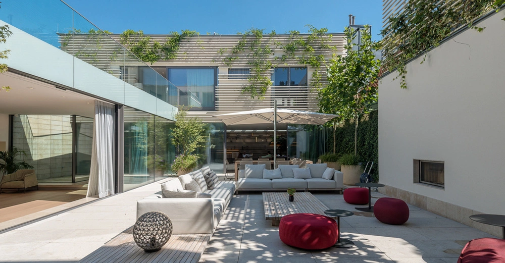 Große, moderne Außenterrasse mit beigen Sofas, roten Hockern und Holztischen vor einem zweistöckigen, begrünten Haus mit großen Fenstern Terrasse umgeben von Pflanzen und Glaswänden in sonniger Umgebung.