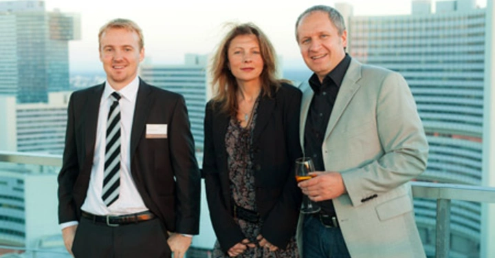 Galerie-Bild 30: Drei Personen stehen auf einer Dachterrasse in einer Großstadt Ein Mann im Anzug links Eine Frau in der Mitte Ein Mann in einem grauen Blazer hält ein Getränk rechts Hochhäuser im Hintergrund