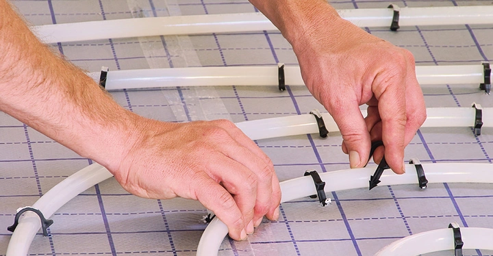 Nahaufnahme der fachgerechten Verlegung einer Fußbodenheizung. Ein Installateur befestigt die Heizungsrohre mit speziellen Clips auf der Systemplatte, um einen optimalen Abstand für die Wärmeverteilung zu gewährleisten.