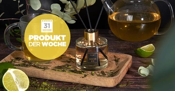 Ein Glas Tee steht auf einem hölzernen Tablett neben einem Diffusor im Vordergrund, mit einem geschnittenen Limette und Teekanne im Hintergrund. Text: Produkt der Woche.