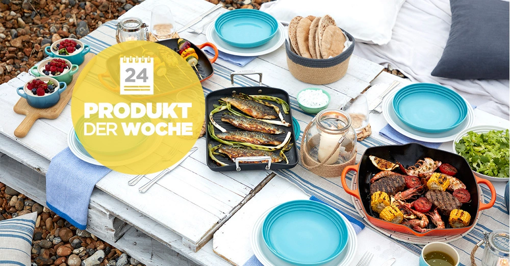 Ein gedeckter Tisch mit blauem Geschirr und verschiedenen Speisen. Grillfisch, Gemüse und Brot werden präsentiert. Draußen auf einem weißen, abgenutzten Holztisch. Text: 24 PRODUKT DER WOCHE.