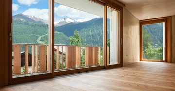 Holzboden eines leeren Raumes großes Fenster auf die Berge und den Wald draussen ein Balkon mit Holzgeländer blauer Himmel mit einigen Wolken der Raum hat beige Wände und Holzeinrahmungen