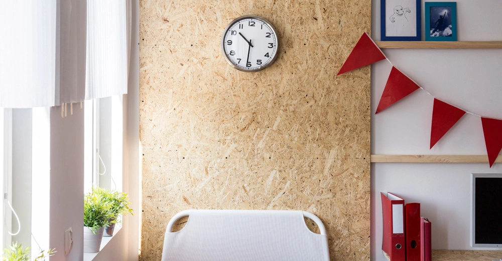 Eine Uhr zeigt 10 Uhr an und hängt an einer Spanplattenwand in einem hellen Raum mit Schreibtisch, roten Aktenordnern, Topfpflanzen und roten Wimpeln an der Wand.